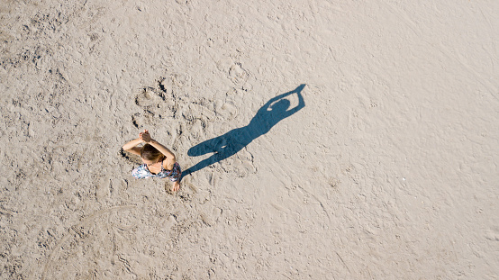 Drone shot of woman exercising on sand, standing on one leg, the other bent inwards, arms raised above head with hands clasped, casting shadow in shape of figure, yoga tree pose practiced outdoors