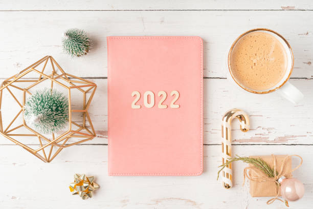 istock Top view Desktop Christmas pink notepad with 2022 letters text. Flat lay on white wooden table background with planner, cup of coffee, candle, toys, Christmas decoration, notebook and stationery 1346326159