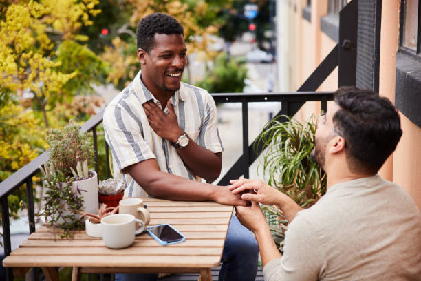 Gay man proposing to his surprised partner on their balcony Young gay man kneeling on one knee and proposing to his surprised partner while sitting together outside on their apartment balcony fiancé stock pictures, royalty-free photos & images