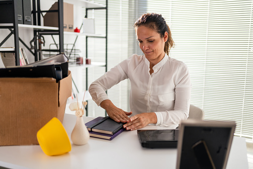 Smiling new female employee unpacking box with belongings at workplace, happy hired office worker newcomer on first working day concept, excited millennial businessman put laptop on desk in coworking