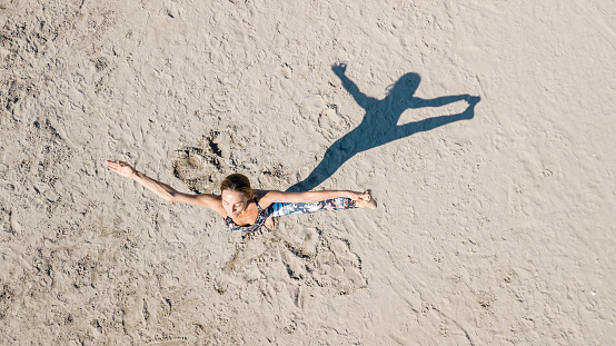 Drone view of woman exercising on the sand standing and holding one leg up with arms outstretched, her shadow in shape of figure she is making, practicing yoga outdoors