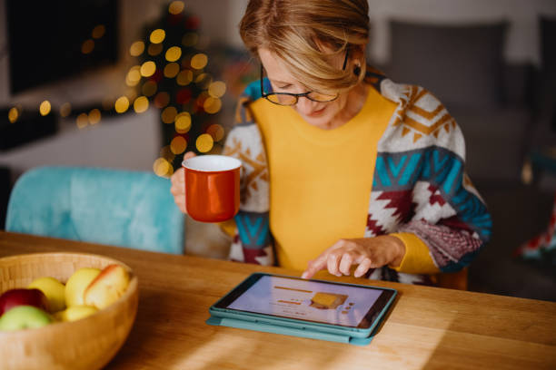 Mature woman using digital tablet for online Christmas shopping Woman enjoying online shopping and using digital tablet and credit card while sitting at dining table home shopping stock pictures, royalty-free photos & images