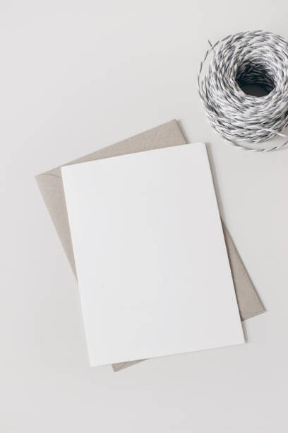 Stationery mockup scene. Blank greeting card, invitation with craft envelope and gray paper rope isolated on white table background. Artistic template, vertical neutral flat lay, top view. No people. escena de maqueta de papelería. tarjeta de felicitación en blanco, invitación con sobre artesanal y cuerda de papel gris aislada sobre fondo de mesa blanca. plantilla artística. colocación plana neutral vertical, vista superior. no hay gente.
