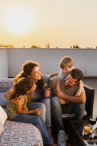 Happy parents and their small kids talking and having fun on a terrace at sunset. Copy space.
