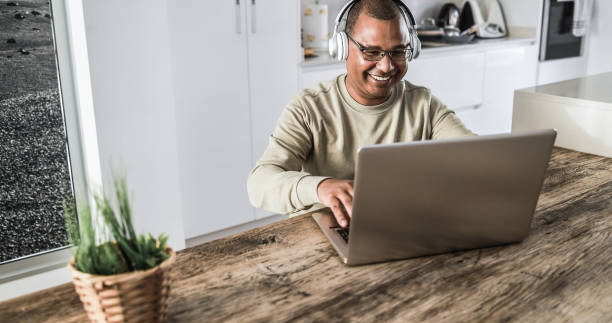Senior african man using laptop computer while wearing headphones at home - Focus on face stock photo