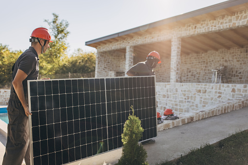Two people, male engineers with protective helmets carrying solar panel. Professional electricians working on installation of solar panels,