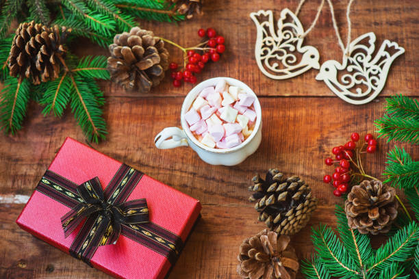 istock Christmas frame. Spruce branches, red berries, bird figures, gift and cup with marshmallows on a wooden background 1346351252