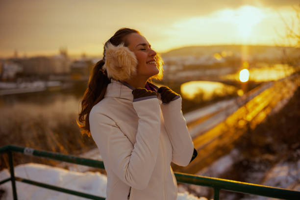 relaxed active woman in white jacket with ear warmer outdoors in the city in winter at sunset.