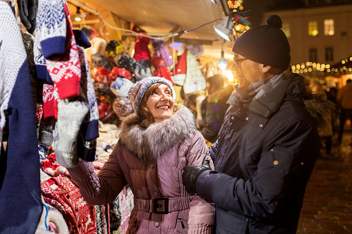 shopping, winter holidays and people concept - happy senior couple at christmas market clothing shop on town hall square in tallinn, estonia