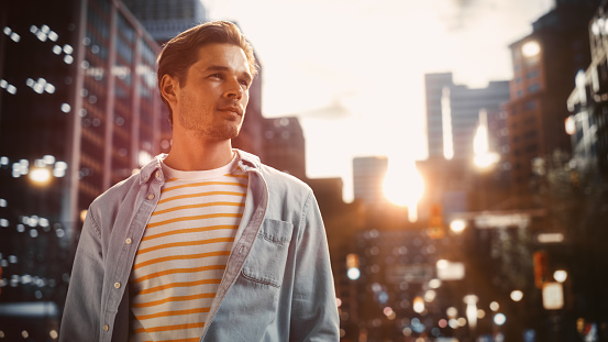 Portrait of a Happy Handsome Young Man in Casual Clothes Standing on the Street at Sunset. Stylish Male Model in Big City Living the Urban Lifestyle. Background with Office Buildings and Billboards.