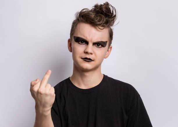 Punk goth-style teen boy Crazy teen boy with spooking make-up making of punk goth dressed in black, showing middle finger on gray background. Problems of transitional age emo boy stock pictures, royalty-free photos & images
