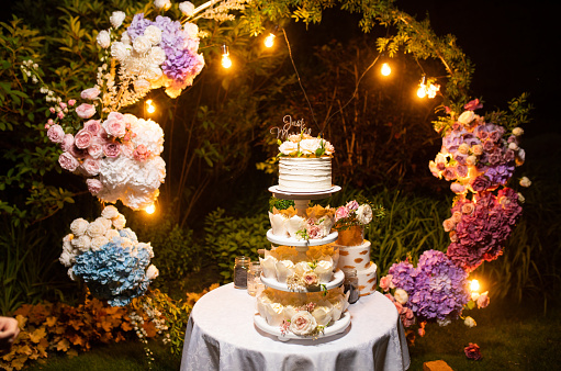 Wedding cake is on table with white tablecloth on the background of wedding arch decorated fresh flowers with glowing garland in garden on lawn.