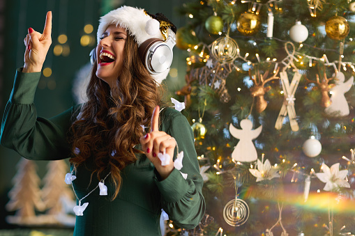 Christmas time. happy stylish woman with Santa hat in green dress listening to the music with headphones and dancing near Christmas tree at modern home.