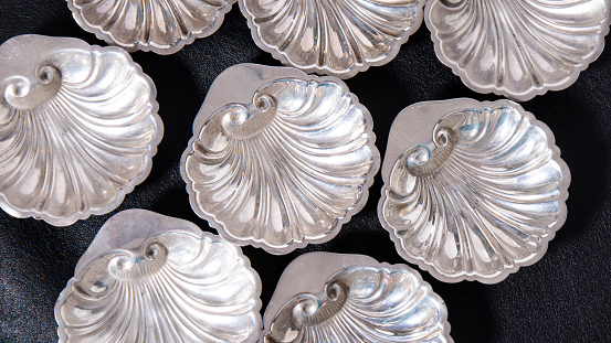 A collection of shiny old silver saucers in the shape of shells on black leather. Shot from above. Beautiful antique silverware.