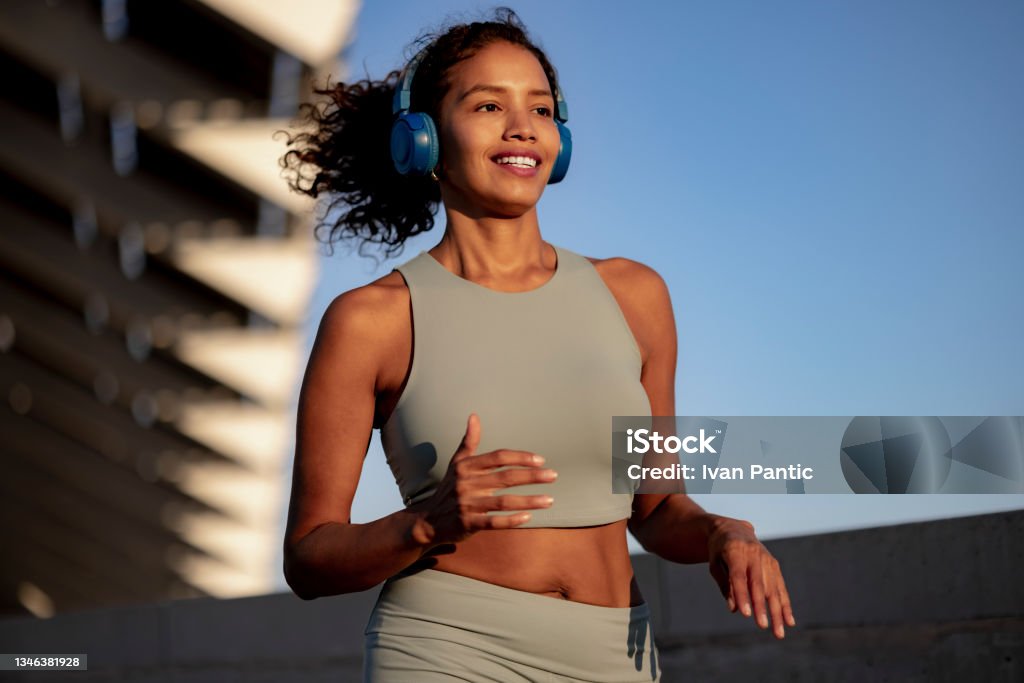 Young Latina sportswoman leading an active lifestyle outdoors Happy fit Latina woman jogging by herself outdoors, following her daily physical activity routine while listening to music on her wireless headphones Running Stock Photo