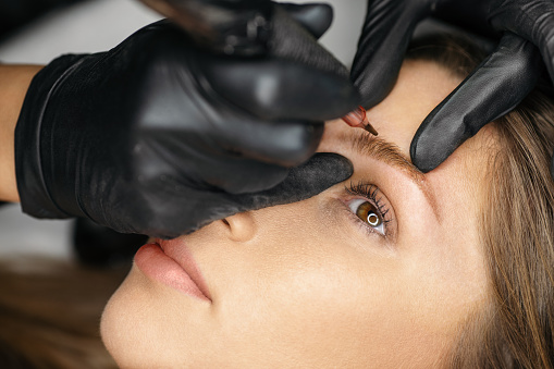 Beautician applying permanent make up microblading technique for eyebrows. She is using device and pen for special tattoo pigmentation and shaping eyebrow