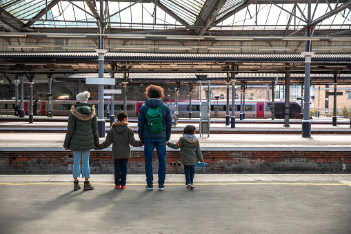 A multiracial family of four, a mother, father and their two little boys on a staycation in the city of Newcastle on a sunny winters day. They are wearing casual, winter clothing and accessories. They are standing on a railway station platform waiting for their train.