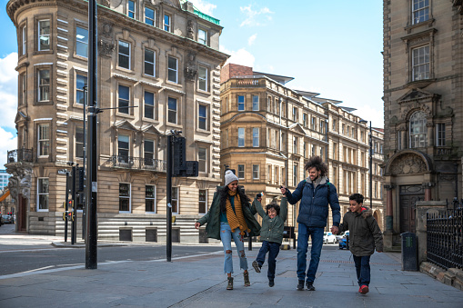 A multiracial family of four, a mother, father and their two little boys on a staycation in the city of Newcastle on a sunny winters day. They are wearing casual, winter clothing and accessories. They are walking down a city street, holding hands and swinging one of the boys in the air.