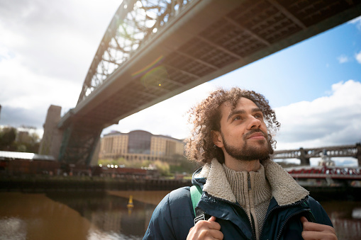 A mixed race man on a staycation in the city of Newcastle on a sunny winters day. He is wearing casual, winter clothing and a backpack. He is standing by the river and taking in the views.