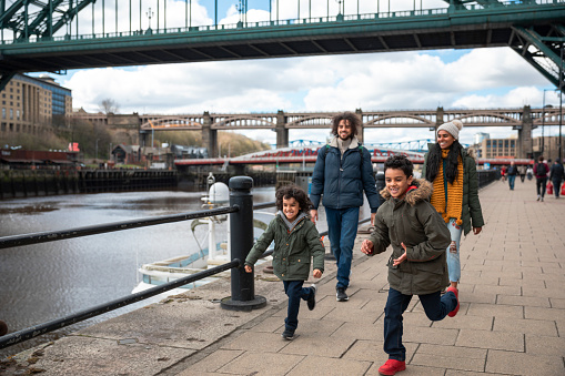 A multiracial family of four, a mother, father and their two little boys on a staycation in the city of Newcastle on a sunny winters day. They are wearing casual, winter clothing and accessories. They are walking down by the river while the two boys run ahead excitedly.