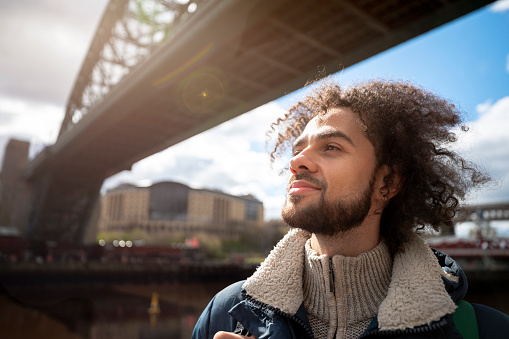 A mixed race man on a staycation in the city of Newcastle on a sunny winters day. He is wearing casual, winter clothing and a backpack. He is standing by the river and taking in the views.