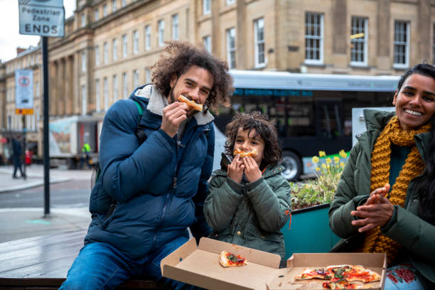 Silly Pizza Face A multiracial family of three, a mother, father and their little boy on a staycation in the city of Newcastle on a sunny winters day. They are wearing casual, winter clothing and accessories. They are sitting and tucking into a takeaway vegan pizza in the city centre. The father and son are messing about and are using the crusts of the pizza as smiles. english cuisine stock pictures, royalty-free photos & images