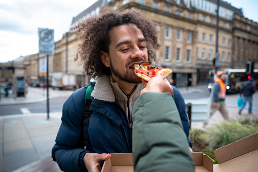 A multiracial couple on a staycation in the city of Newcastle on a sunny winters day. They are wearing casual, winter clothing and accessories. It is a point of view shot from an unrecognisable woman feeding her partner a slice of pizza in the city centre.