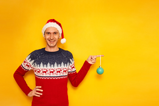 happy, cheerful excited man in Christmas sweater with reindeer, red Santa Claus hat holds tree toy in hands and points finger empty area for advertising. isolated yellow background with space for text. High quality photo