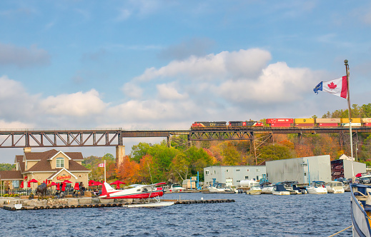 Parry Sound, Ontario/Canada: October 10, 2021: Scenic autumn view of waterfront including marina with boats, restaurant, Trans Canada Railway with CN cargo train, sightseeing seaplane and tourists