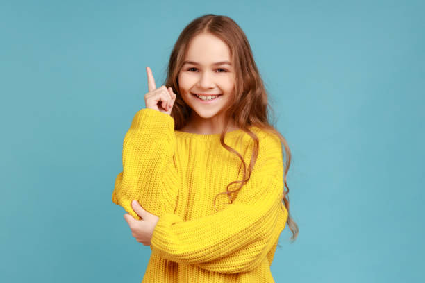Portrait of little girl points finger up, looking inspired by genius thought, showing good idea sign Portrait of little girl points finger up, looking inspired by genius thought, showing good idea sign, wearing yellow casual style sweater. Indoor studio shot isolated on blue background. 9 stock pictures, royalty-free photos & images