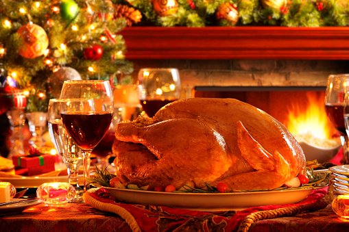 A Christmas turkey sits on a dinner table in front of a roaring fireplace and a Christmas tree.