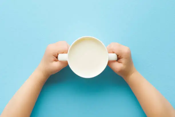 Toddler hands holding cup of white fresh milk on light blue table background. Pastel color. Closeup. Point of view shot. Daily healthy drink. Top down view.