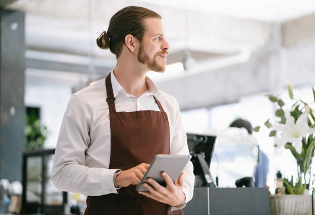 istock Portrait of bearded hipster barista using digital tablet and looking away while standing at trendy coffee shop. Happy barista man are taking orders from online customers. Startup small business owner. 1346399763