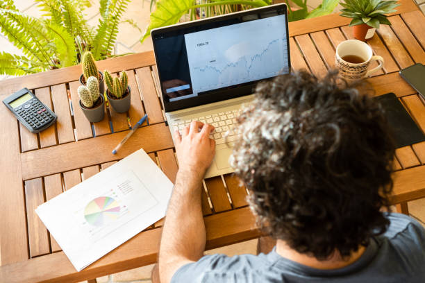 jovem usando computador para analisar gráficos e fazer investimentos em estoque enquanto bebe uma xícara de café em home office cheio de plantas. - unrecognized person - fotografias e filmes do acervo