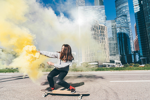 Young woman rolling longboard with color smoke grenade at the city. Side view of female practicing skating.