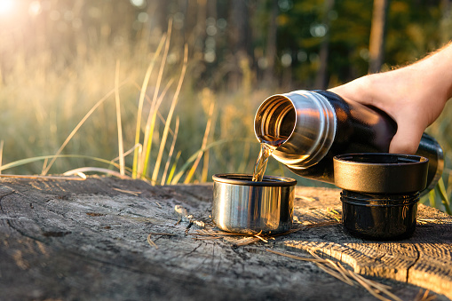 Tourist pouring hot tea out of a thermos flask into two cups standing on a tree stump at sunset. Hiking, traveling and active lifestyle concept