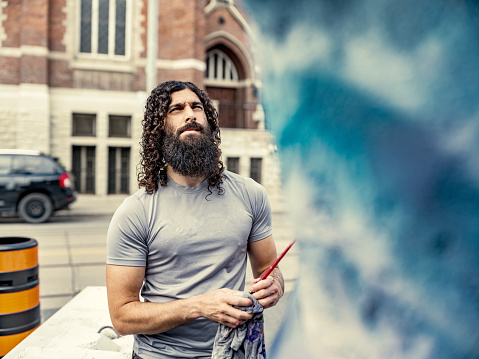Young artist painting finish on his street sculpture. He has long black curly hair and beard, dressed in t-shirt and jeans. Exterior of city street sidewalk  in downtown district of North American city.