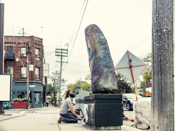 Young artist painting finish on his street sculpture. He has long black curly hair and beard, dressed in t-shirt and jeans. Exterior of city street sidewalk  in downtown district of North American city.