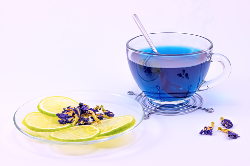 Cup of Anchan or Butterfly pea tea with dry flowers of Clitoria. Thai Blue tea in a transparent glass cup with slices of lime and dry flowers isolated on white background.