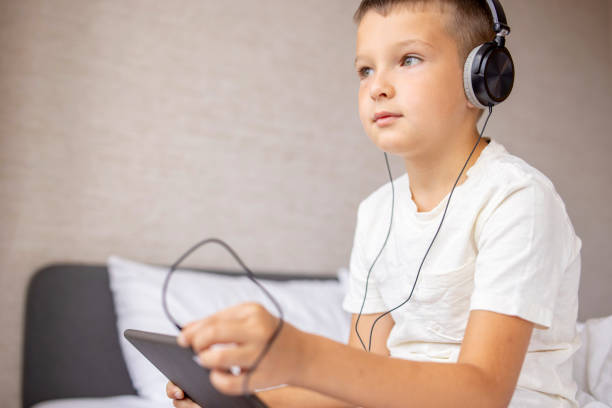 istock Smiling happy kid caucasian boy in white T-shirt siting on bed, using tablet computer with headphones in bed at home 1346420561