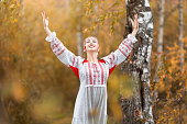 Portrait of young beautiful smiling woman in folk traditional slavic clothes waving hands  and asking for blessing from spirits at nature in autumn forest. Pagan slavs rituals and traditions concept