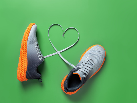 Shoelace heart symbol between gray textile sneakers with grooved orange sole on grassy green background. Laced up stylish mesh fabric sport shoes with heart shape symbol for active lifestyle and fitness. Top view.
