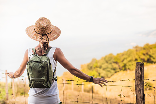a woman hiker with hat and backpack, contemplates a landscape leaning against a fence. she is on her back looking at the horizon.