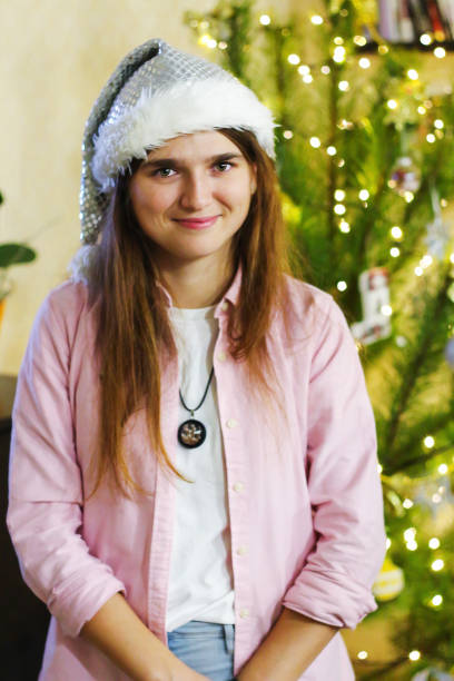 白いサンタの帽子とピンクのシャツを着て笑顔の青写真女子学生20-25年の肖像画。背景にクリスマスツリーと屋内ポーズ。グリンチのように見えます。クリスマス。選択的フォーカス - 20 25 years color image contemporary domestic life ストックフォトと画像