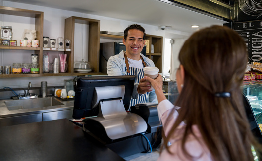 Happy Latin American barista working at a cafe and handling a cup of coffee to a woman at the counter