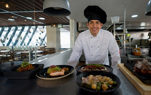 Happy Latin American chef presenting his plates in the kitchen of a restaurant for service and looking at the camera smiling - food and drink concepts