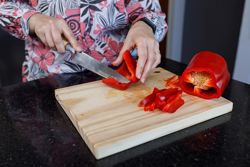 A Woman's hand with a wedding ring, cutting a red bell bell pepper on a wooden board.