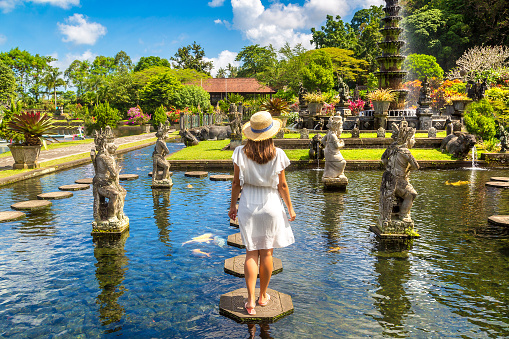 Woman traveler wearing white dress and straw hat at  Taman Tirtagangga temple on Bali, Indonesia in a sunny day