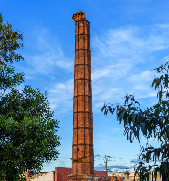 A brick construction factory chimney is in disrepair, framed by green leaves and against a blue sky background