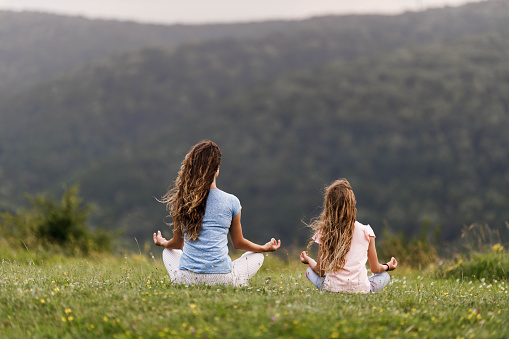 Rear view of mother and daughter doing Yoga relaxation exercises in Lotus position at the park. Copy space.
